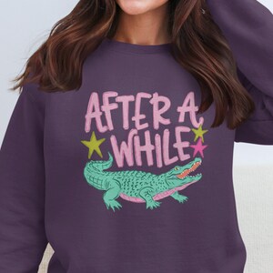 May include: A purple sweatshirt with the words "AFTER A WHILE" in pink, with a green alligator illustration. Yellow and pink stars are also included in the design. The sweatshirt is a crew neck style.