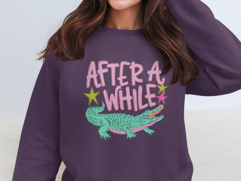 May include: A purple sweatshirt with the words "AFTER A WHILE" in pink, with a green alligator illustration. Yellow and pink stars are also included in the design. The sweatshirt is a crew neck style.