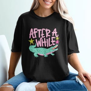 May include: Black t-shirt with the text "AFTER A WHILE" in pink, with a green alligator illustration. The alligator has pink accents and is surrounded by a yellow star and a pink star.