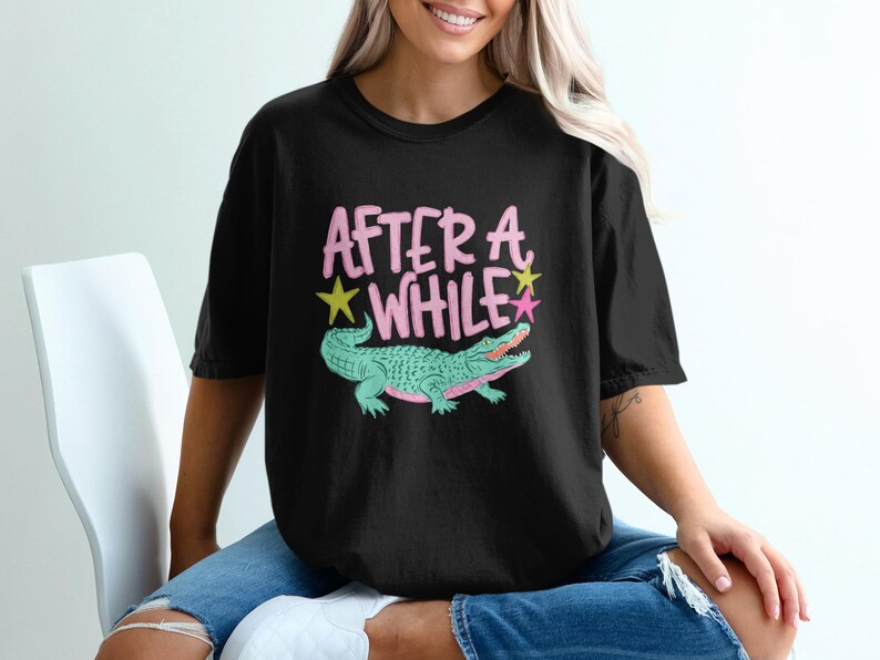 May include: Black t-shirt with the text "AFTER A WHILE" in pink, with a green alligator illustration. The alligator has pink accents and is surrounded by a yellow star and a pink star.