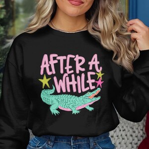 May include: Black sweatshirt with the text "AFTER A WHILE" in pink, with a green alligator illustration and yellow and pink stars. The sweatshirt is worn by a person.