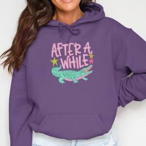 May include: Purple hoodie with a kangaroo pocket and drawstring hood. The front features the text "AFTER A WHILE" in pink, with a teal alligator illustration and star accents. A casual, comfortable garment.