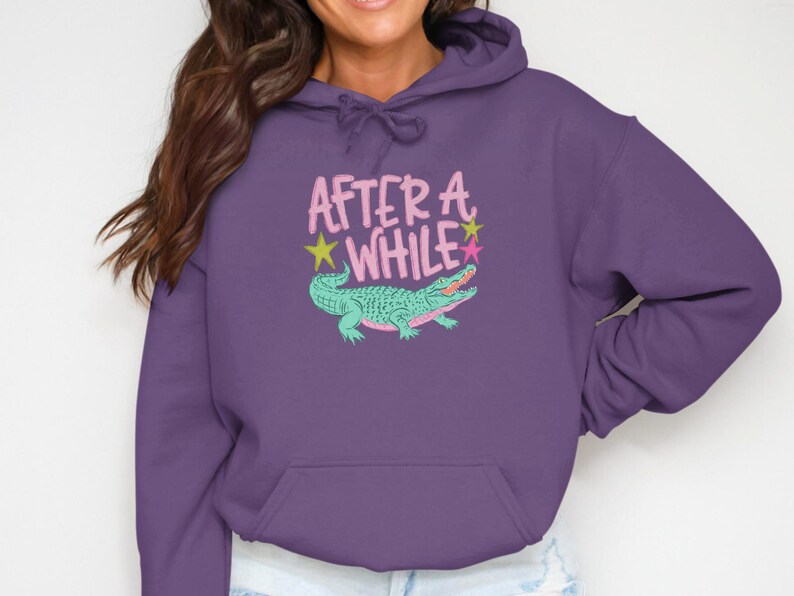 May include: Purple hoodie with a kangaroo pocket and drawstring hood. The front features the text "AFTER A WHILE" in pink, with a teal alligator illustration and star accents. A casual, comfortable garment.
