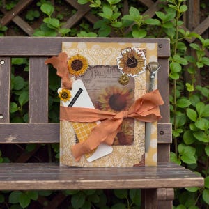 Könnte beinhalten: Ein dekoratives Notizbuch mit Sonnenblumen-Motiv, mit einem Sonnenblumen-Bild im Vintage-Stil, verziert mit Stoff- und Papier-Sonnenblumen und einem Band. Ein Stift ist darin versteckt. Das Notizbuch liegt auf einer Holzbank.