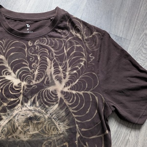 Puede incluir: Una camiseta de manga corta de color marrón oscuro con un diseño gráfico blanqueado de un ojo grande rodeado de patrones orgánicos y arremolinados similares a telarañas. La etiqueta interior muestra el texto Tu y la talla M. Cuello redondo.