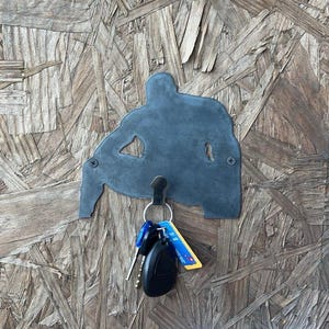 May include: A dark grey metal wall hook shaped like a silhouette of a person sitting down. The hook is secured with two visible screws and holds a metal ring with a black car key fob and a blue and yellow plastic tag attached to the key ring.