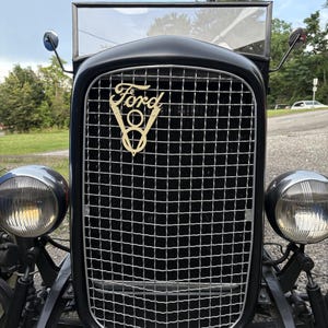 May include: Close-up of a vintage black Ford car. The car features a chrome grill, two round headlights, and the Ford logo in gold. The windshield is clear, and the car appears to be in good condition.