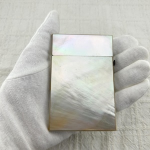 May include: A rectangular cigarette case featuring a polished mother-of-pearl inlay on the front surface. The case has a hinged lid design with a metallic frame and a smooth, iridescent finish that displays subtle white, cream, and soft pastel tones.