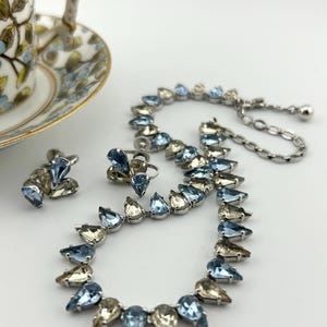 Op de afbeelding: Een vintage sieradenset met een ketting, oorbellen en een ring. De set is versierd met druppelvormige, lichtblauwe en heldere kristallen, gezet in zilverkleurig metaal. Een bloemenkop en schotel zijn op de achtergrond.