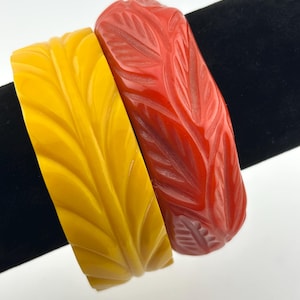 May include: Two wide bangle bracelets featuring a carved leaf pattern. One bangle is a solid mustard yellow color, and the other is a solid deep red color. Both bracelets have a smooth, glossy finish and a repeating textured design across the surface.