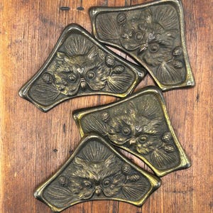May include: Five antique bronze-toned metal decorative trays. Each tray features a detailed owl design with a textured surface and a gold-colored border. The trays are arranged on a wooden surface.