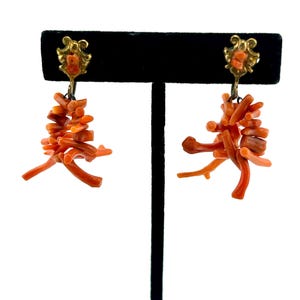 May include: A pair of coral earrings. Each earring features a cluster of orange coral pieces suspended from a gold-colored filigree top. The coral pieces are arranged to resemble small branches. The earrings are displayed on a black stand against a white background.