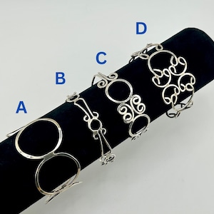 Könnte beinhalten: Vier silberfarbene Metallarmreifen, präsentiert auf einer schwarzen Samtoberfläche. Jedes Armband hat ein einzigartiges filigranes Design mit kreisförmigen und dekorativen Elementen. Die Armreifen werden vor einem hellen Hintergrund präsentiert.