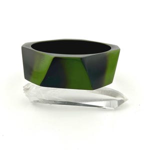 May include: A hexagonal bangle bracelet featuring a smooth, polished surface with a marbled pattern of deep forest green and black tones, resting on a clear, pointed, prismatic crystal shard against a plain white background.