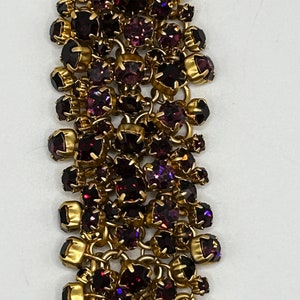 Op de afbeelding: Een gouden kettingarmband met meerdere rijen paarse strass steentjes. De armband is gemaakt van metaal en heeft een delicaat, vrouwelijk ontwerp.