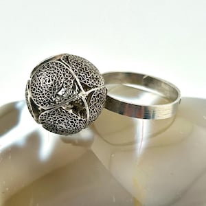 May include: A silver ring featuring a large, spherical filigree ornament composed of intricate, swirling wire patterns. The band of the ring is a smooth, plain silver metal. The spherical centerpiece is constructed from multiple curved sections.
