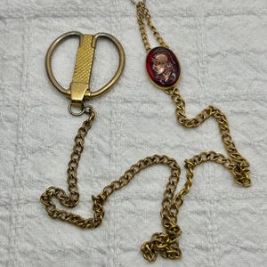 May include: A gold chain with a gold and silver clasp and a red cameo with a gold profile of a person wearing a helmet.