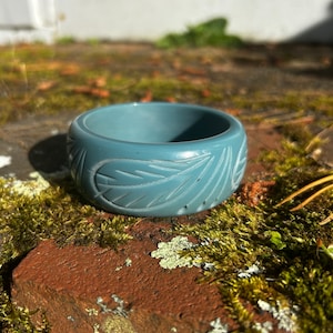 Puede incluir: Un brazalete azul en forma de bangle con un diseño de hojas talladas. El brazalete está hecho de un material liso y pulido y tiene una forma ligeramente redondeada.