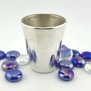 May include: A small, silver-coloured cup with a textured surface. The cup is surrounded by blue and iridescent glass pebbles. The cup has a simple, elegant design, suitable for various uses.