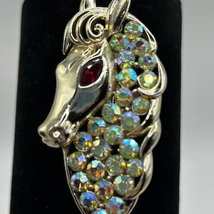 Könnte beinhalten: Eine silberfarbene Pferde-Brosche mit einem roten Strassstein-Auge und schillernden Strasssteinen. Die Mähne und der Schweif des Pferdes bestehen aus Strasssteinen.