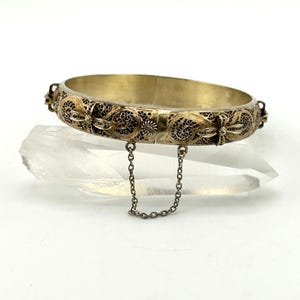 May include: A gold-coloured hinged bangle bracelet with detailed filigree and floral patterns. The bracelet has a safety chain and clasp. The filigree has dark accents, and the bracelet is set on a clear, crystalline surface.