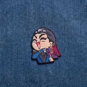 May include: An embroidered patch featuring a stylised chibi character with dark hair, a blue outfit, and a purple accent, laughing with eyes closed. The patch is applied to a textured blue denim fabric background with a visible horizontal seam.