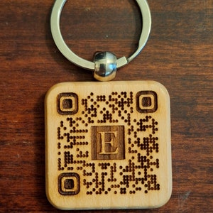 Op de afbeelding: Een houten sleutelhanger met een lasergegraveerde QR-code en de letter "E" in het midden. De sleutelhanger is bevestigd aan een zilveren metalen ring.