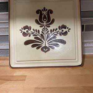 May include: Square ceramic serving dish with a cream-colored background and a brown floral design. The dish has a brown border and is sitting on a wooden surface. The floral design is centered on the dish.