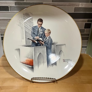May include: A decorative plate with a gold rim, depicting a man and a boy reading in a church. The illustration is detailed and signed by Norman Rockwell. The plate's design features soft, muted colours, creating a classic and elegant aesthetic.