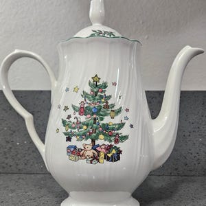 May include: A white porcelain teapot with a scalloped rim and a matching lid. The body features a colorful illustration of a decorated Christmas tree with ornaments, a star topper, and wrapped gifts with a teddy bear at the base of the tree.