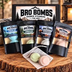 May include: A black cardboard box labeled Bro Bombs Shower Steamers for Men containing four black pouches with labels Fresh AF, Lumberjack, Barbershop, and Morning Wood. Two round shower steamer tablets in light green and tan sit on a small mesh bag.