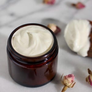 May include: A small, brown glass jar filled with white cream. The cream has a smooth, swirled texture. A wooden spoon with cream on it is in the background, along with dried rose buds. The jar is on a white marble surface.