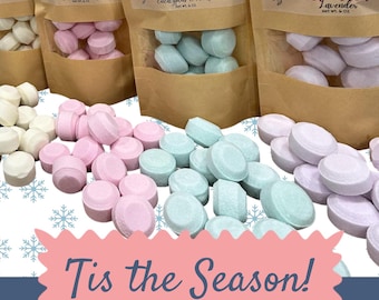 May include: A set of 12 shower steamers in four different scents: Mood Boost, Sinus Sooth, Sweet Dream, and Lemon & Lime. The steamers are packaged in clear plastic bags with brown paper labels. The labels feature a mermaid logo and the scent name. The text "Tis the Season!" is printed on a pink and blue background with snowflakes. The text "12 steamers Net wt. 7 oz" is printed below the text "Tis the Season!"
