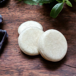 May include: Three white round soap bars on a wooden surface. The soap bars are stacked on top of each other.