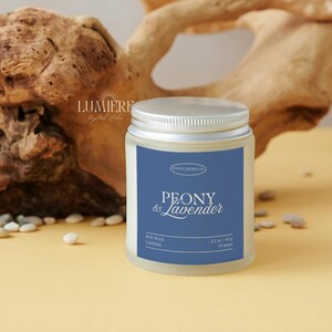 K&ouml;nnte beinhalten: Ein zylindrisches Kerzenglas mit silbernem Deckel und blauem Etikett. Auf dem Etikett steht "PEONY & Lavender" und "Soy Wax Candle". Das Glas enth&auml;lt 60 g Wachs und hat eine Brenndauer von 11 Stunden. Im Hintergrund Treibholz und kleine Steine.