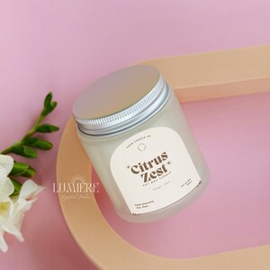 May include: A white soy wax candle in a glass jar with a silver lid. The label reads "Citrus Zest Soy Wax Candle" with the text "Orange & Basil" underneath. The candle is sitting on a light brown surface.