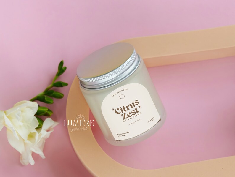 May include: A white soy wax candle in a glass jar with a silver lid. The label reads "Citrus Zest Soy Wax Candle" with the text "Orange & Basil" underneath. The candle is sitting on a light brown surface.