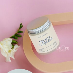 K&ouml;nnte beinhalten: Eine Sojawachskerze in einem cremefarbenen Glas mit silbernem Deckel, beschriftet mit "Peony & Lavender". Die Kerze liegt auf einem pfirsichfarbenen Tablett mit wei&szlig;en Blumen und gr&uuml;nen Bl&auml;ttern vor rosa Hintergrund. 60g.