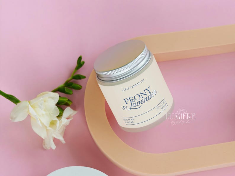 K&ouml;nnte beinhalten: Eine Sojawachskerze in einem cremefarbenen Glas mit silbernem Deckel, beschriftet mit "Peony & Lavender". Die Kerze liegt auf einem pfirsichfarbenen Tablett mit wei&szlig;en Blumen und gr&uuml;nen Bl&auml;ttern vor rosa Hintergrund. 60g.