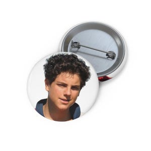 May include: Two white round badges. One badge features a portrait of a person with dark curly hair, wearing a blue and red collared shirt. The second badge is partially visible, showing the pin back.