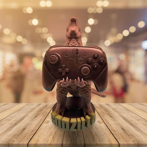 May include: A brown dinosaur-shaped Nintendo Switch controller holder. The dinosaur is holding a Switch controller with its claws. The base of the holder has the word "GODZILLA" in green letters. The controller is dark brown.
