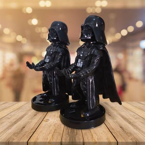 May include: Two black Darth Vader figurines with outstretched hands, standing on round bases. The figurines have glossy black helmets, capes, and armor. The background is blurred, with a wooden table in the foreground.