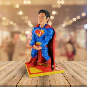 May include: A Superman figurine in a classic pose, featuring a blue suit with the iconic red and yellow chest emblem, a red cape, and red boots. The figurine stands on a yellow and red base.