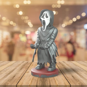 May include: A gray figurine of the Ghostface character from the Scream horror film franchise. The figure is wearing a hooded robe and a white mask with a screaming expression. It stands on a red circular base.