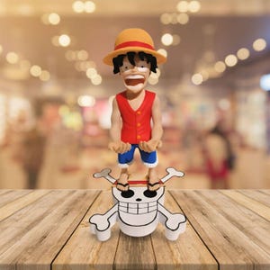 May include: A figurine of a cartoon character wearing a straw hat, red vest, and blue shorts, standing on a white skull and crossbones. The figure has a wide-open mouth and is set on a wooden surface.