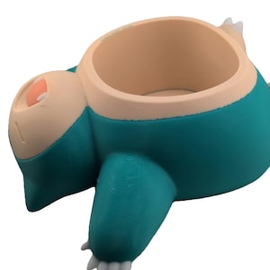 May include: A teal and beige Snorlax planter, a Pokémon character, with a large opening for plants. The planter has a round body, small arms and legs, and a sleeping expression. The planter is made of a hard plastic material.