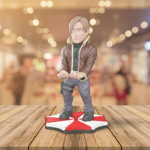 May include: A figurine of a character from the Resident Evil video game series. The figure is wearing a brown jacket, blue pants, and black boots. It stands on a red and white base with a black border. The background is blurred.