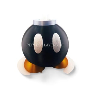 May include: A black and white 3D printed bob-omb figurine with a silver top. The bob-omb has two white eyes and the text "PERFECT LAYERS 3D" is printed on the side. The bob-omb is standing on two gold and white bases.