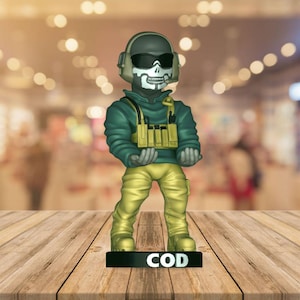 May include: A figurine of a character from the video game Call of Duty. The figure is wearing a green hooded jacket, yellow pants, and a skull mask with sunglasses. The figure is standing on a black base with the letters "COD" in white.
