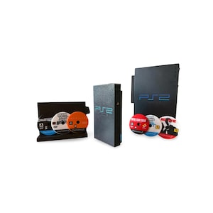 May include: A black PlayStation 2 console with the PS2 logo in blue, accompanied by several game discs. The console is displayed with a vertical stand and a separate disc holder, showcasing the games.